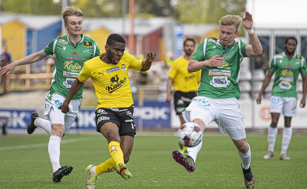 Uzochukwu Henry Onuorah, wearing a yellow jersey and black shorts, kicks a ball past a defender wearing a green jersey and white shorts/